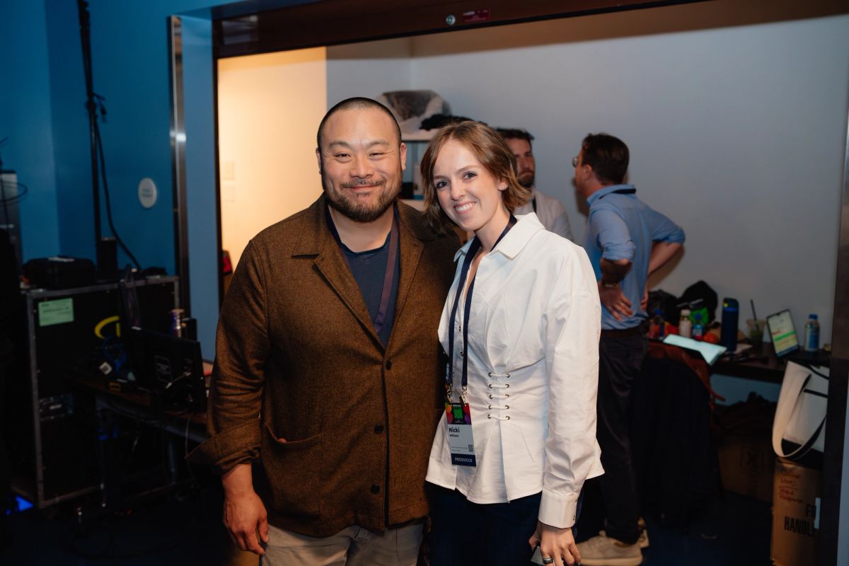 Two conference attendees with lanyards pose together in a venue with filming equipment and blue walls