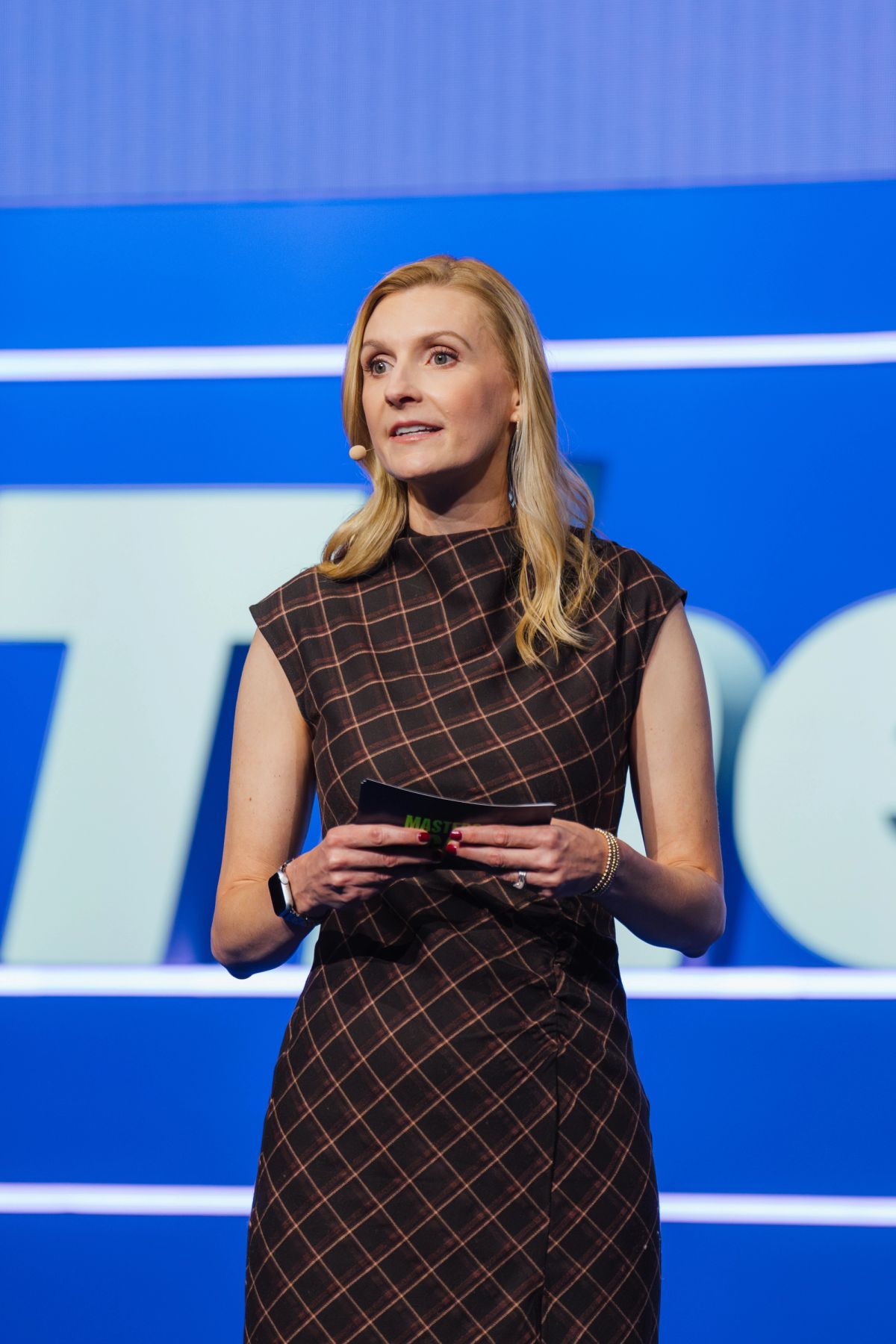 Speaker in dark checkered dress holding device on stage with blue backdrop at conference event.
