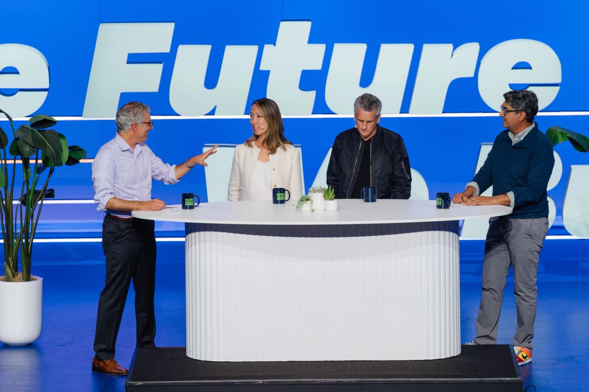 Four people on stage at conference panel with white curved desk and blue backdrop showing "Future" text
