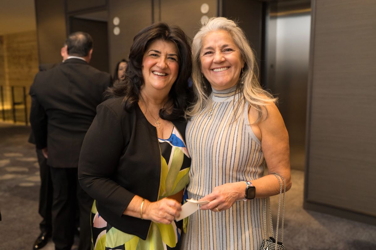 Two women smiling together at an indoor event, one wearing a black blazer and colorful dress, the other in a.