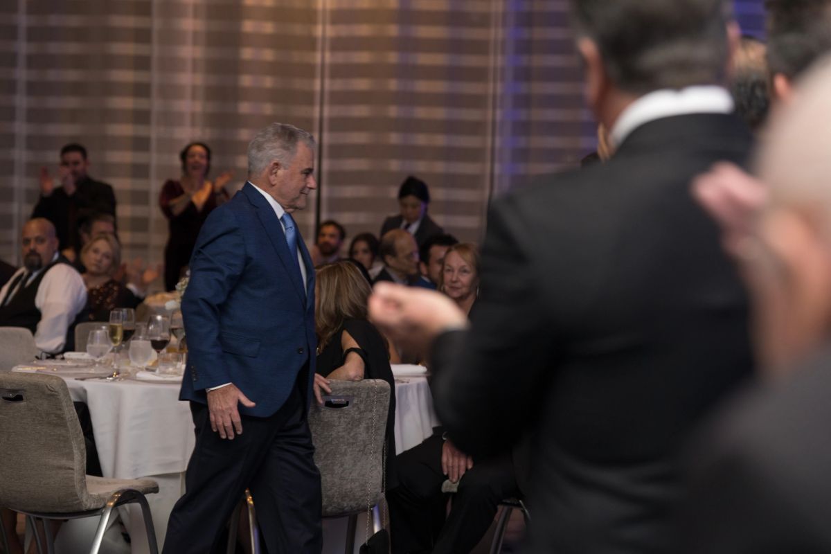 Man in blue suit standing at formal dinner event with seated guests at white-clothed tables