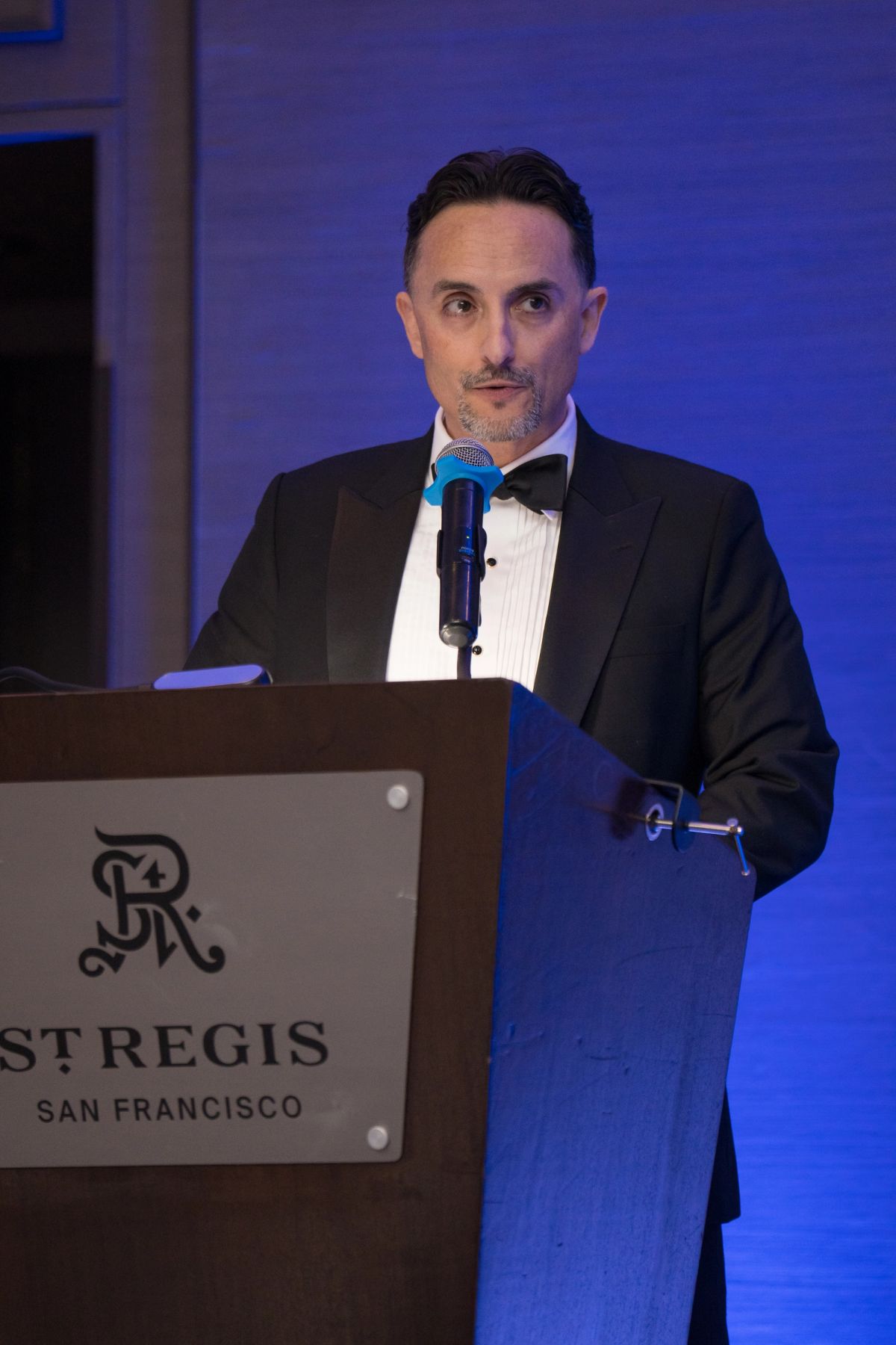 Speaker in black tuxedo presenting at podium with microphone at St. Regis San Francisco gala event