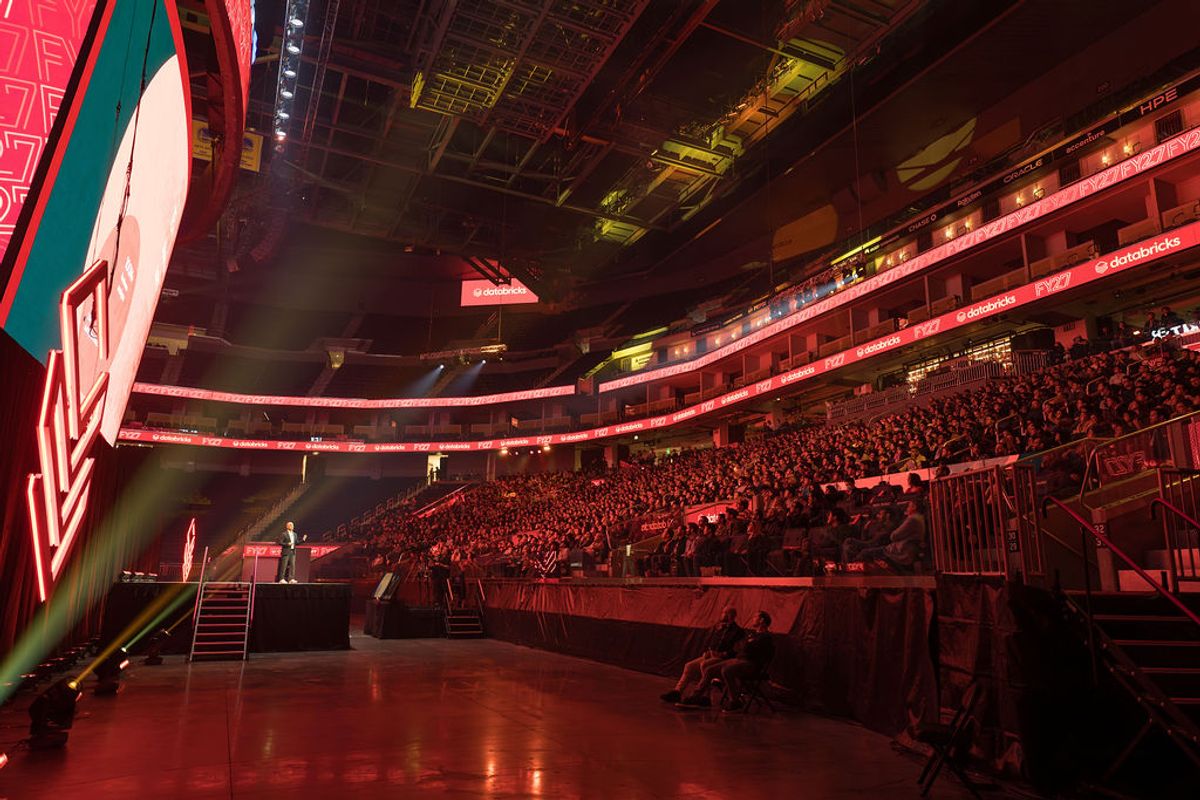 Arena interior with red lighting, packed crowd in tiered seating, and stage area with speakers visible