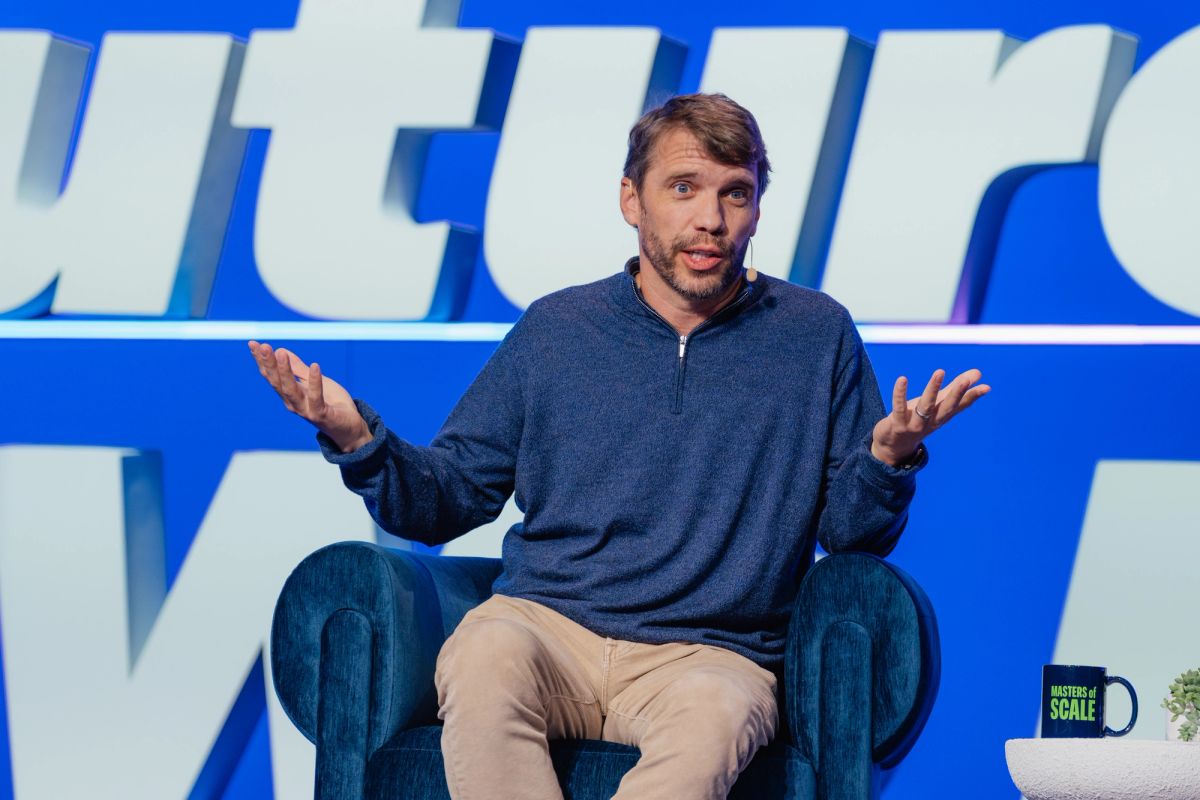 Speaker in blue sweater gesturing while seated in armchair on stage with blue backdrop at conference