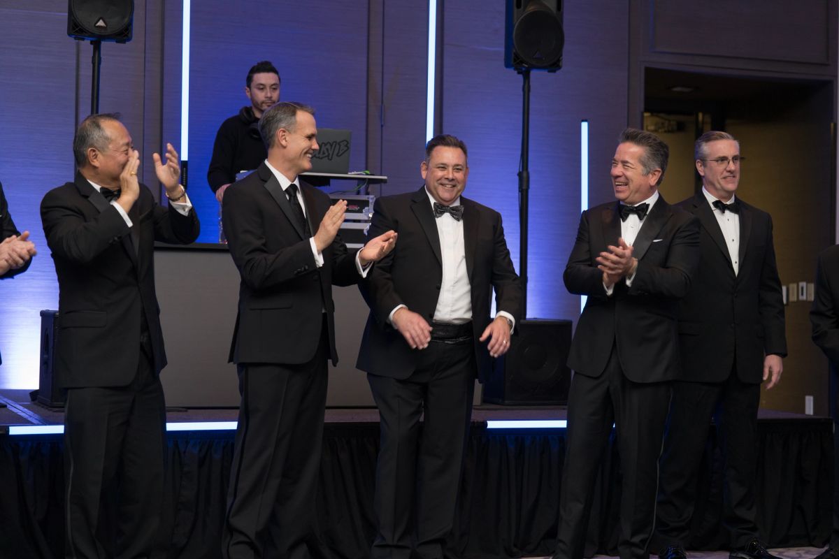 Five men in formal black tuxedos applauding on stage at an upscale gala event with blue lighting.
