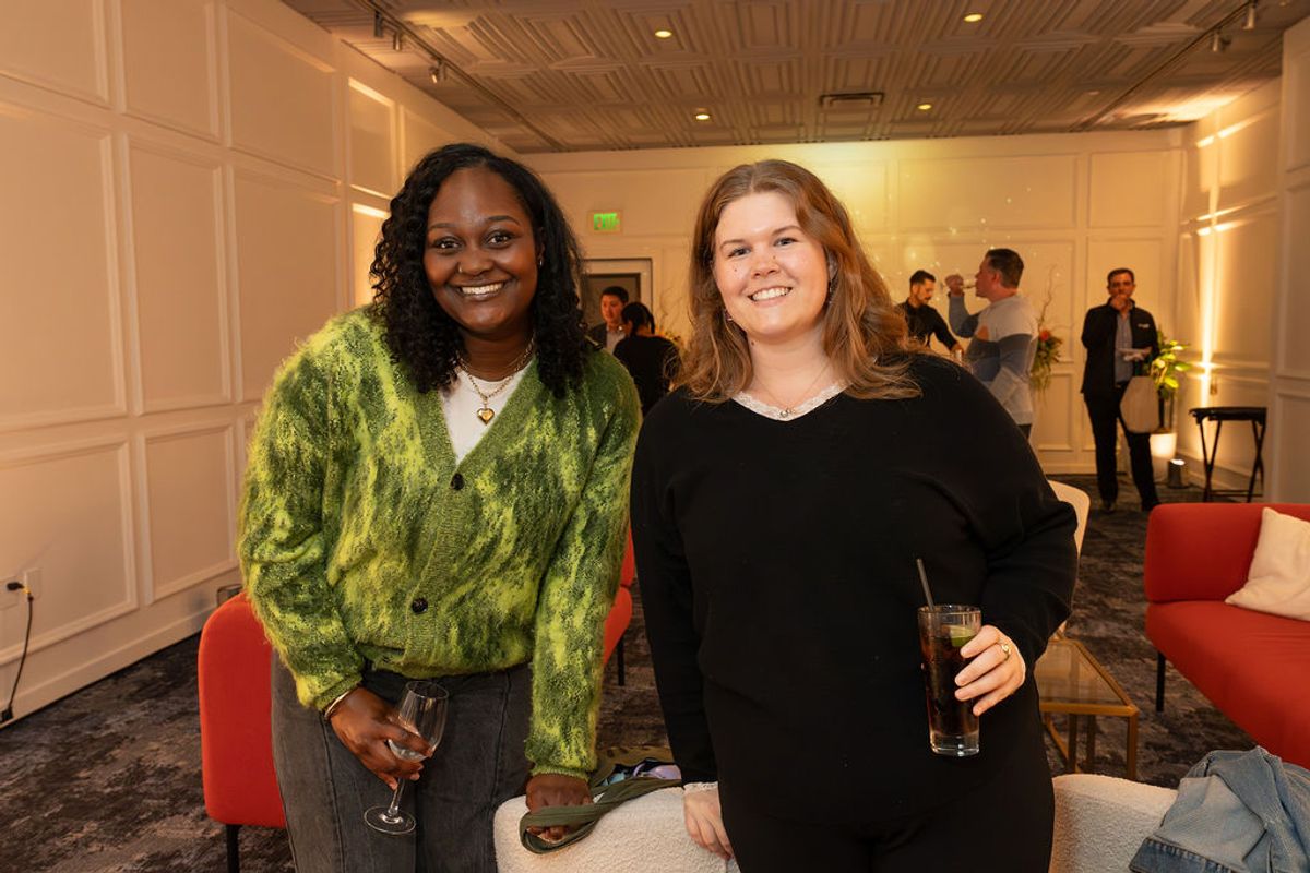 Two women networking at a reception, one in a green sweater and one in black, holding drinks in an elegant.