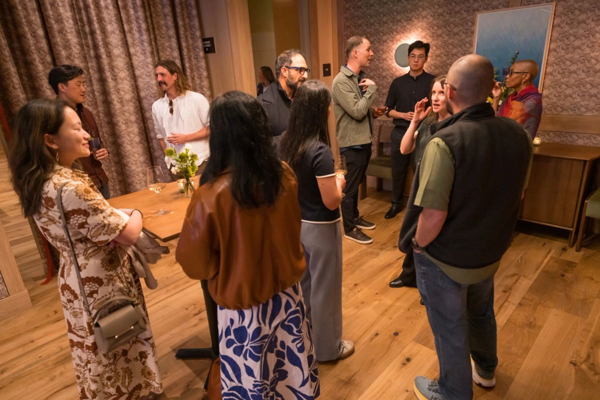 Group of people standing and mingling in a room with wooden floors, curtains, and casual conversation.