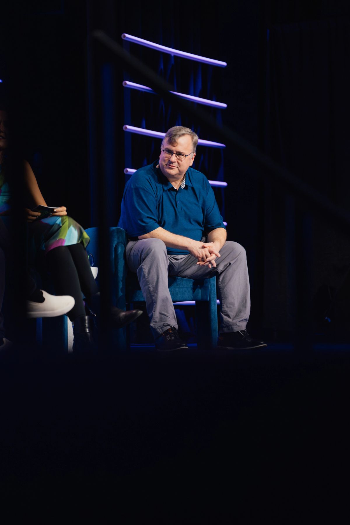 Speaker in blue shirt sits on stage chair during conference presentation with blue lighting