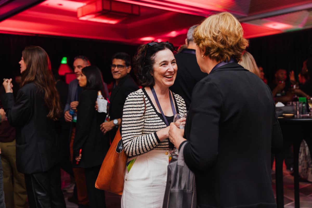 Two women networking and conversing at an indoor conference reception with red lighting and other attendees.