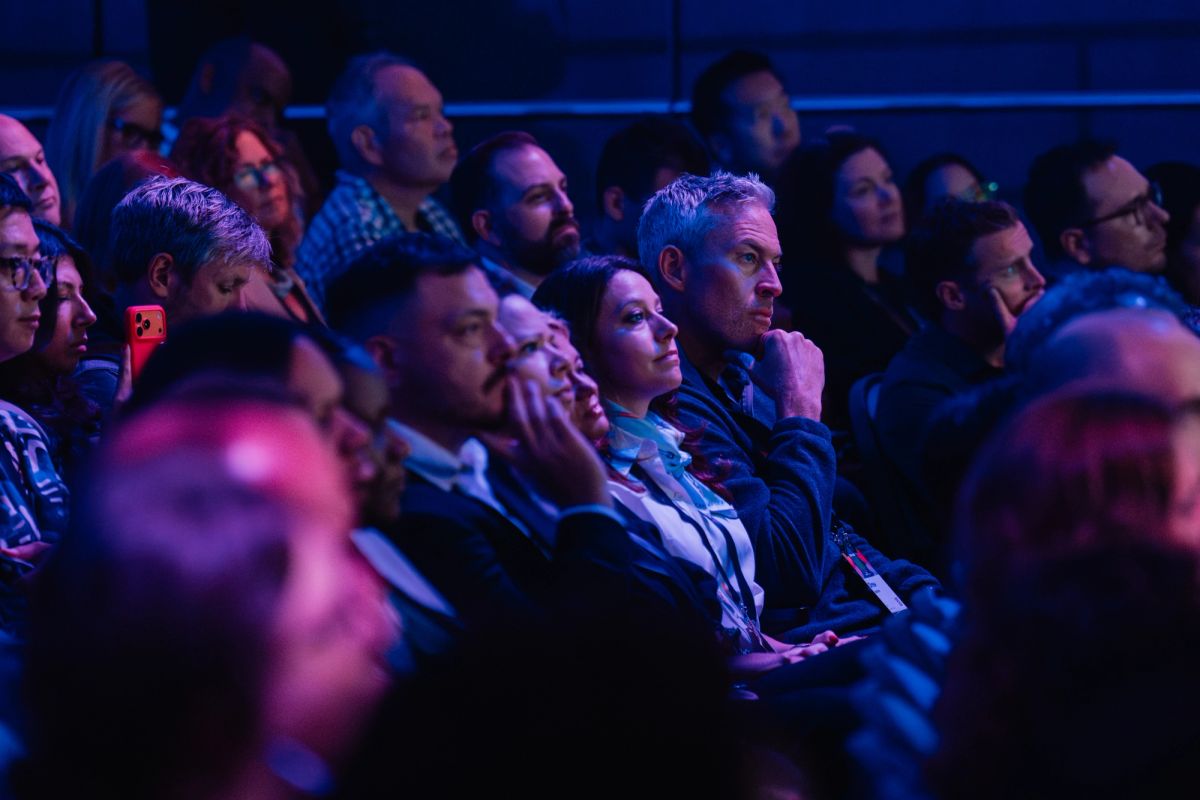 Audience members seated in theater-style seating listen attentively during a conference presentation in dim.