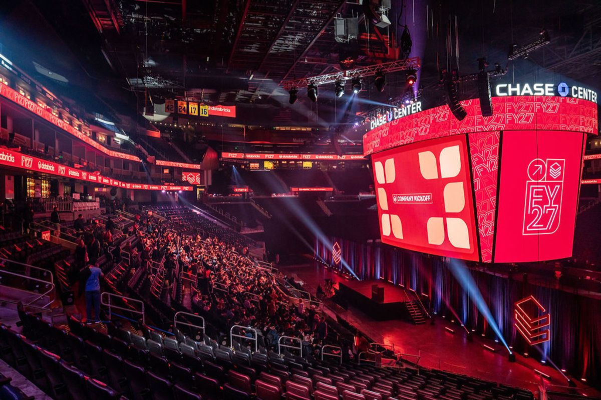 Conference attendees in a large arena with red lighting and screens displaying presentation content