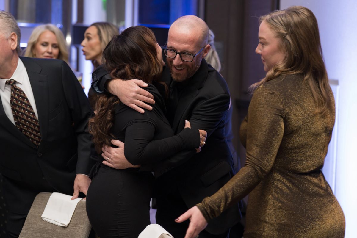 Two people embrace warmly while others look on at a formal indoor gathering