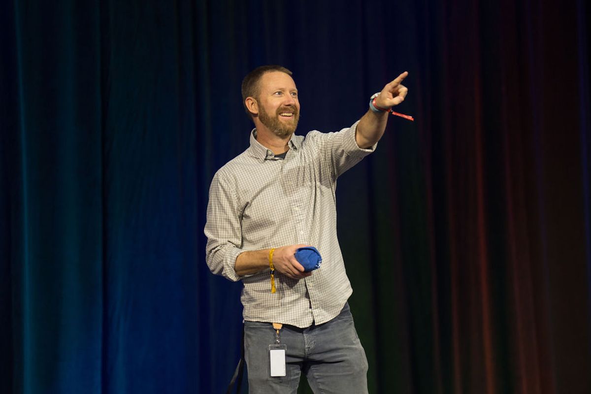 Speaker presenting on stage with blue backdrop, pointing while holding presentation remote