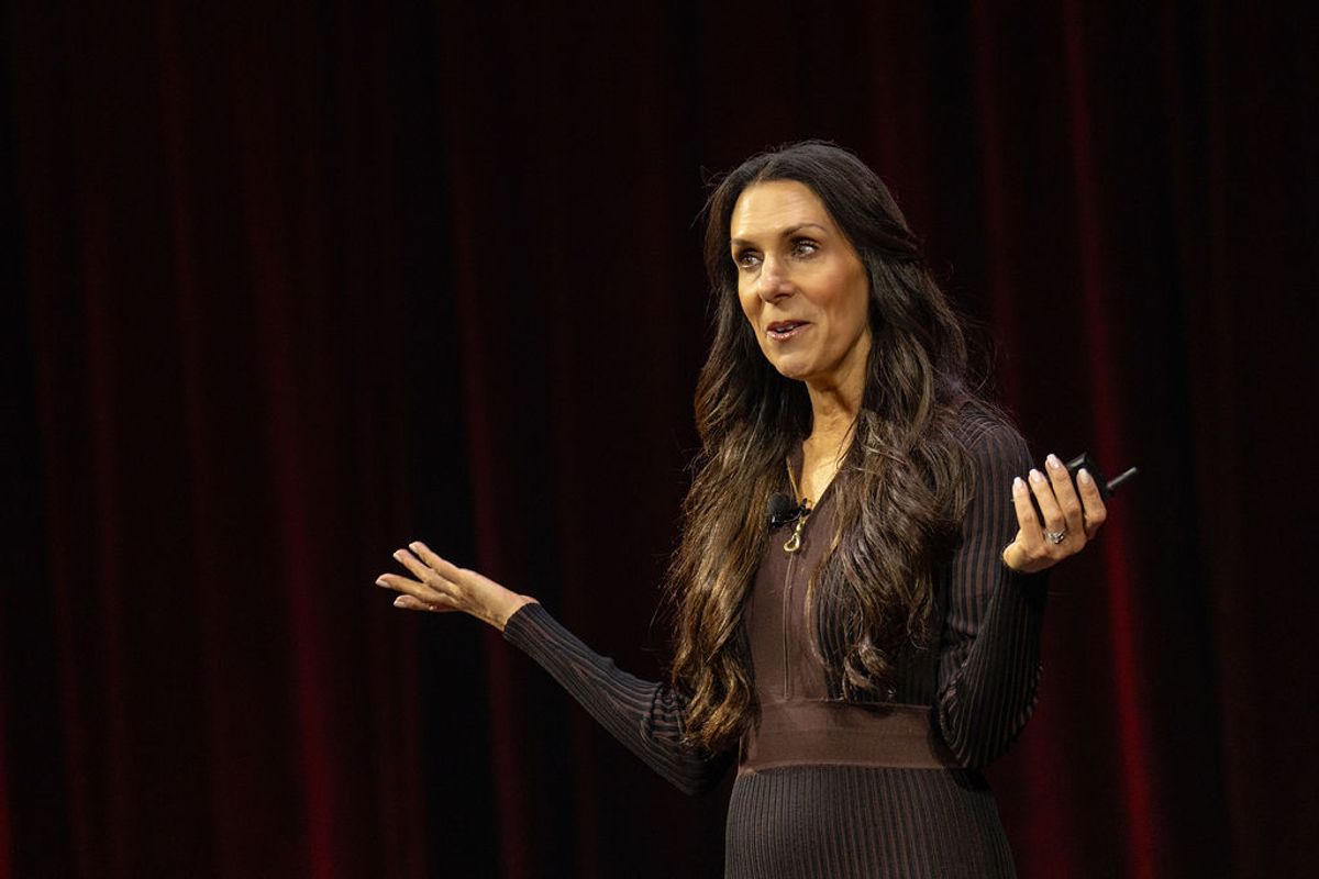 Speaker presenting on stage with microphone, gesturing with both hands against dark backdrop with red curtains