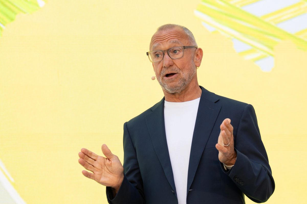 Male speaker gesturing with both hands, wearing blazer and earpiece mic, yellow backdrop behind him.