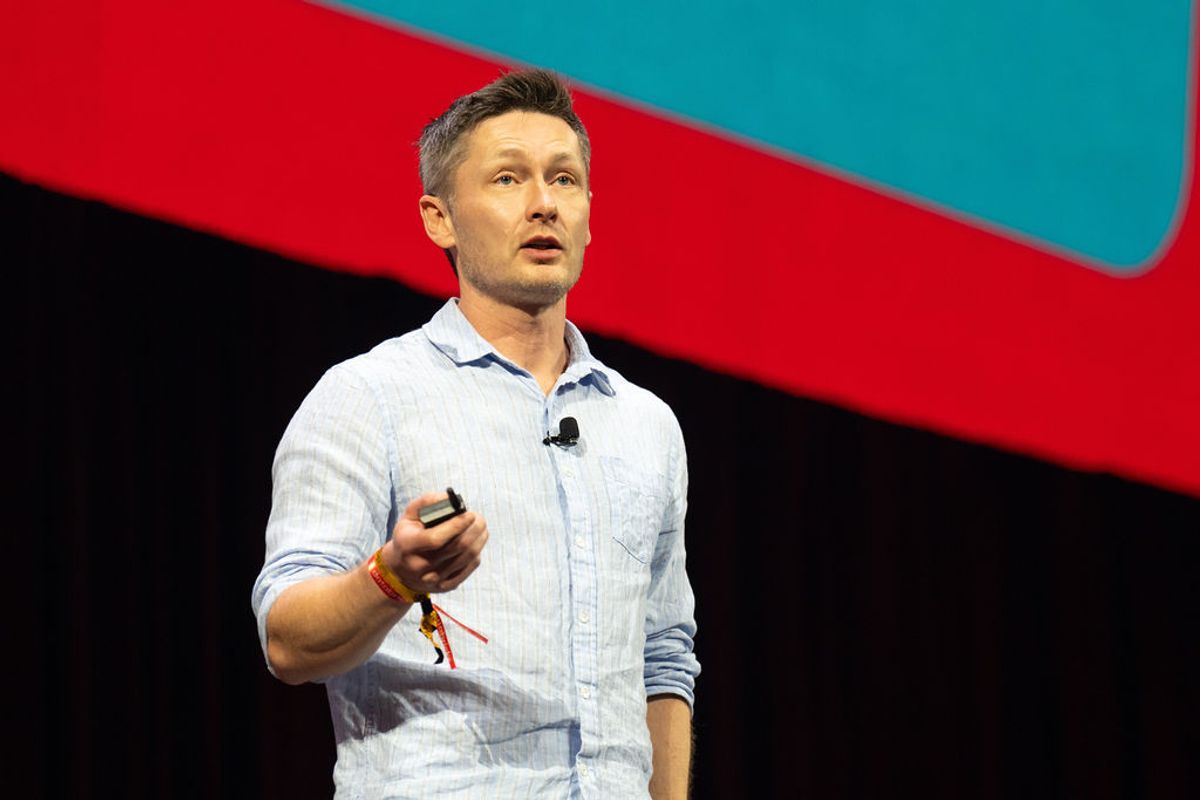 Speaker presenting on stage with microphone at conference, wearing light blue shirt against red backdrop