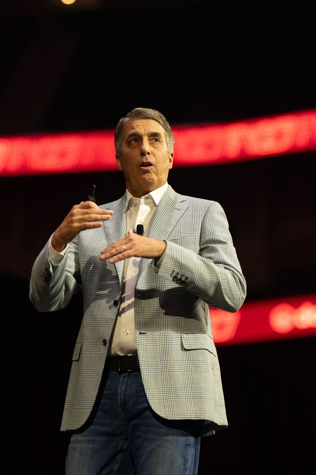 Speaker in gray suit jacket gesturing while presenting on stage with red lighting in background
