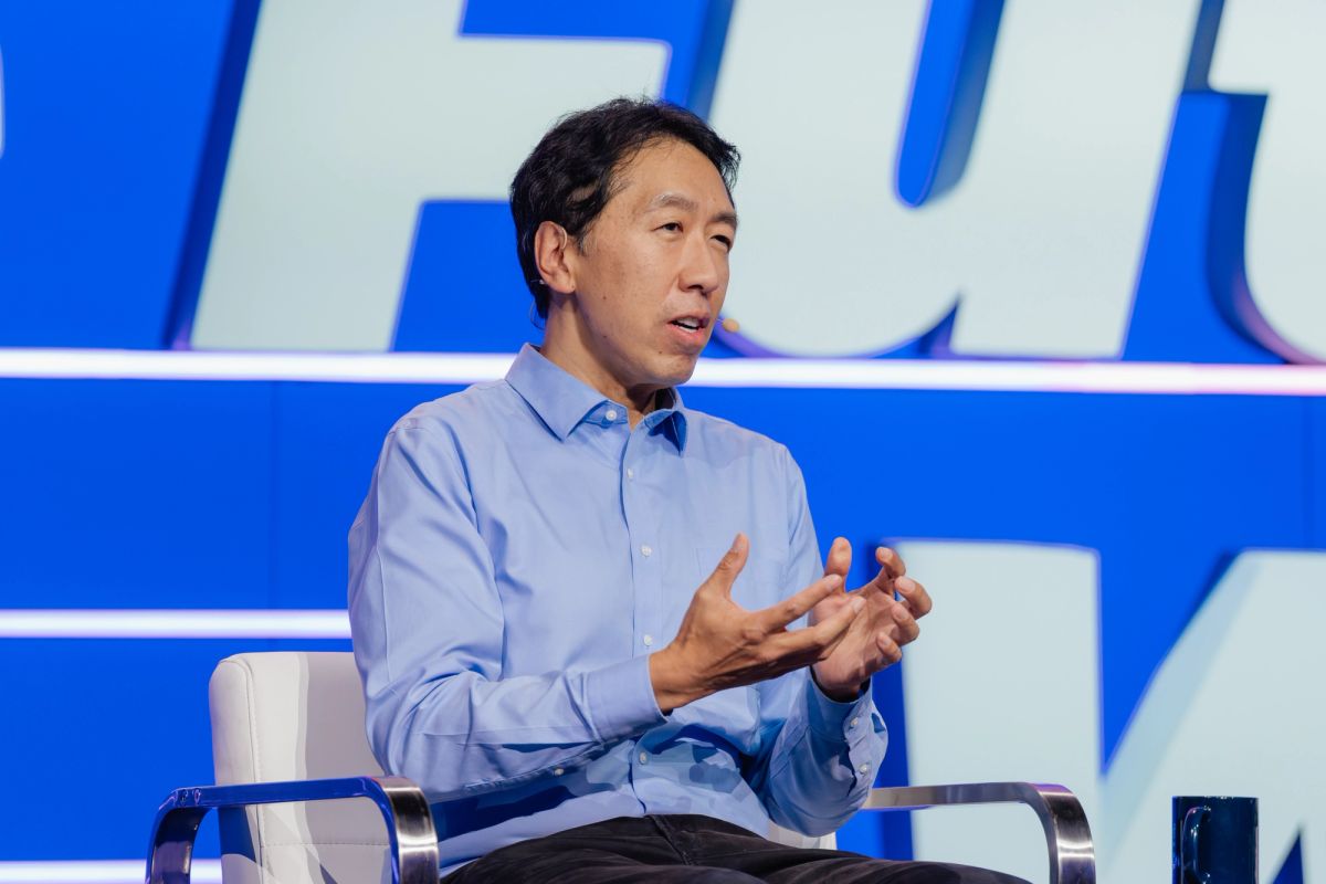 Speaker in blue shirt gesturing while seated in white chair at conference with blue backdrop
