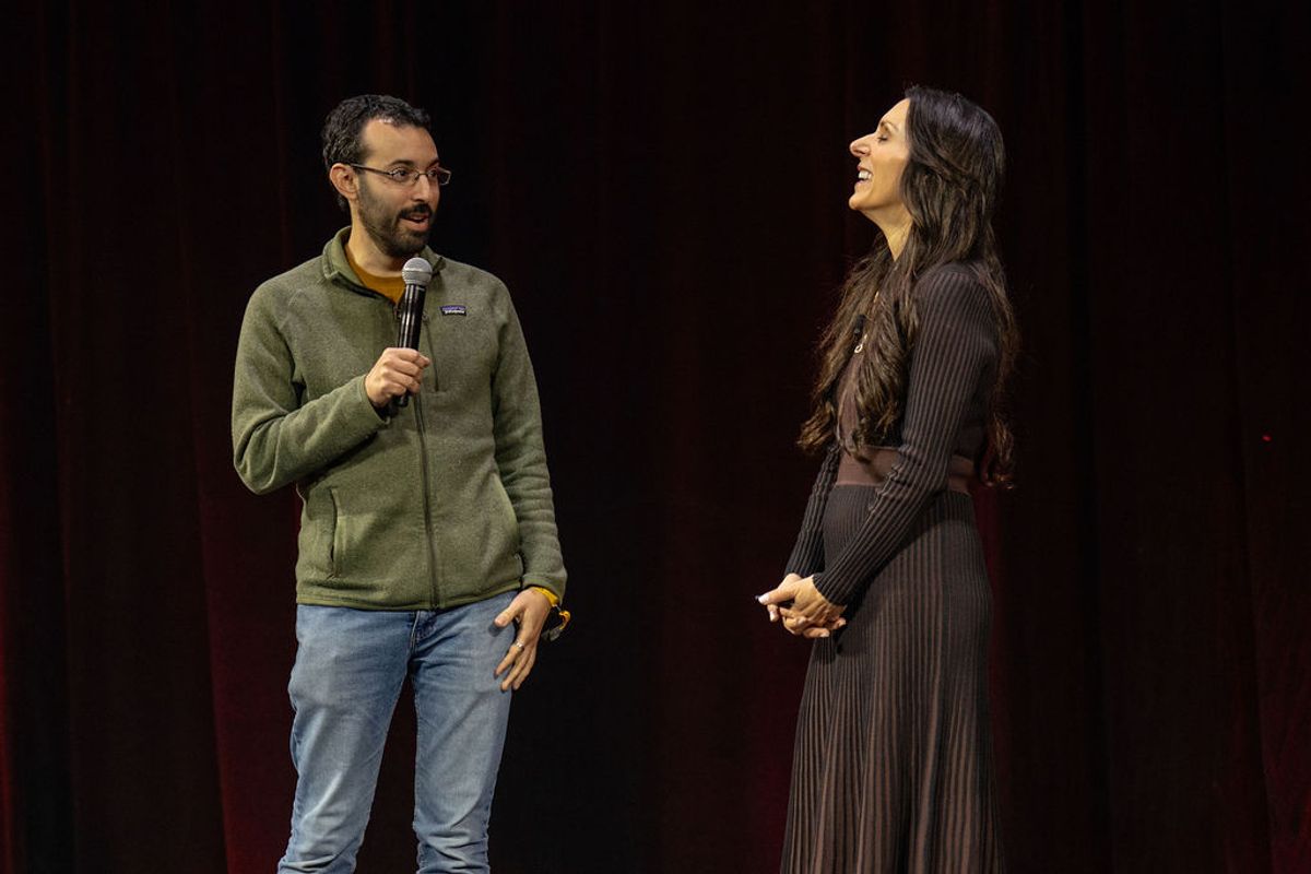 Two speakers on stage, one holding microphone in green sweater, other in black dress, both smiling