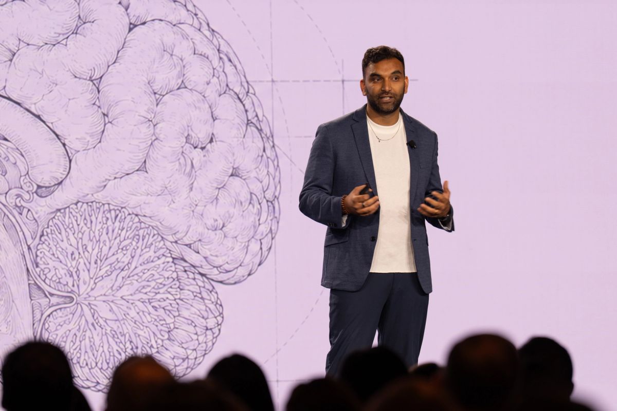 Speaker presenting on stage before large brain illustration backdrop, audience silhouettes in foreground.
