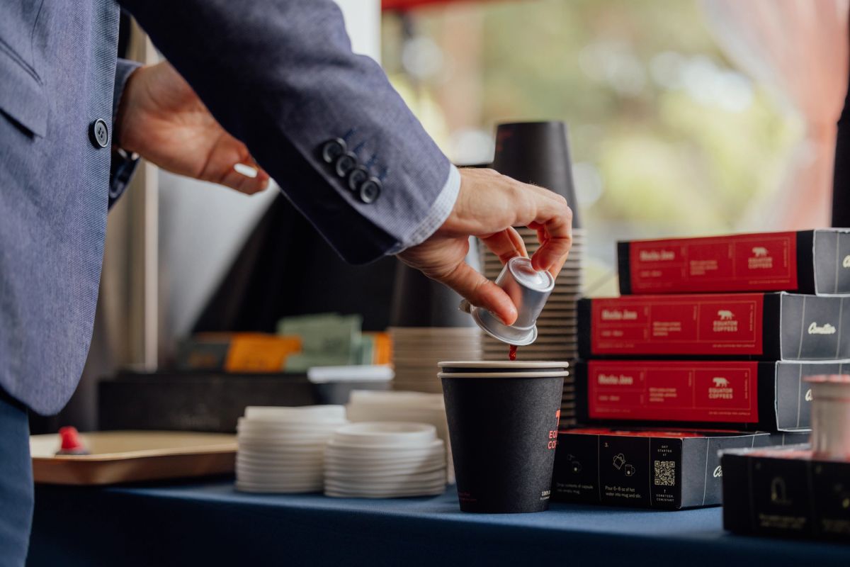 Person in suit pouring from small container into cup at coffee station with stacked boxes and plates