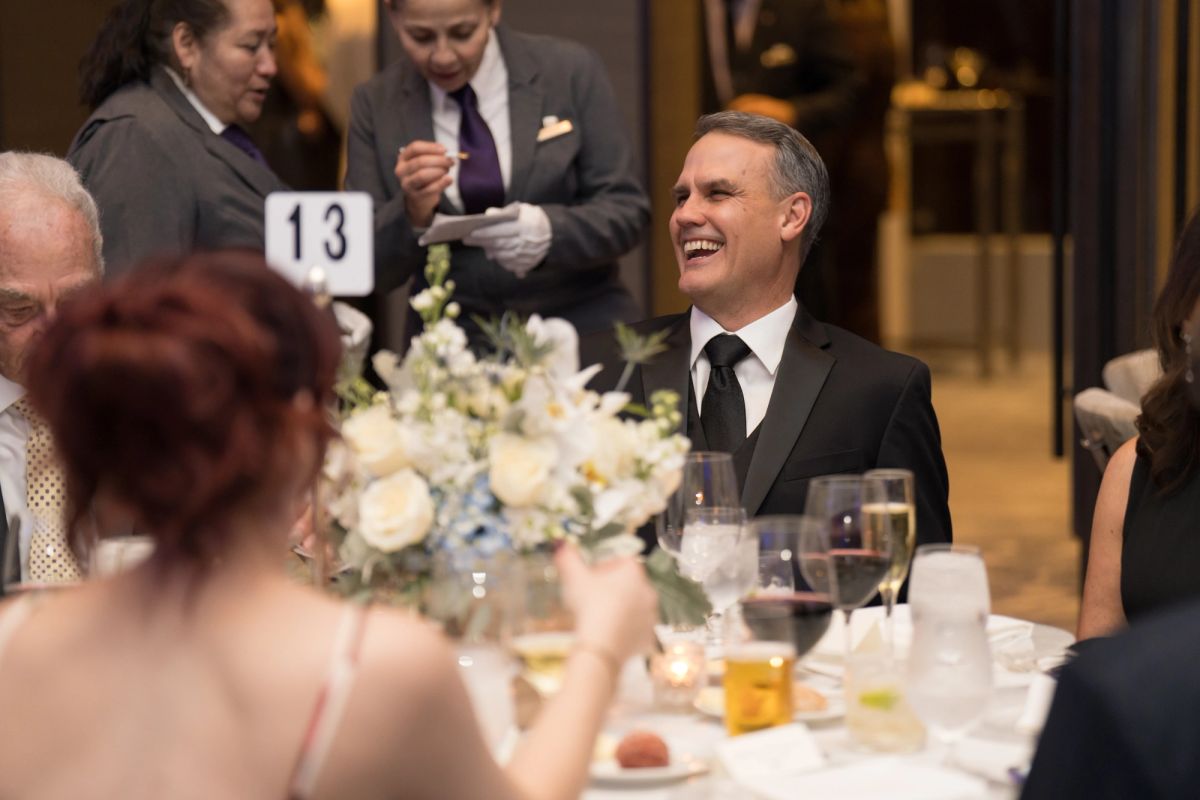 Smiling man in tuxedo at formal dinner table with white floral centerpiece and table number 13 visible