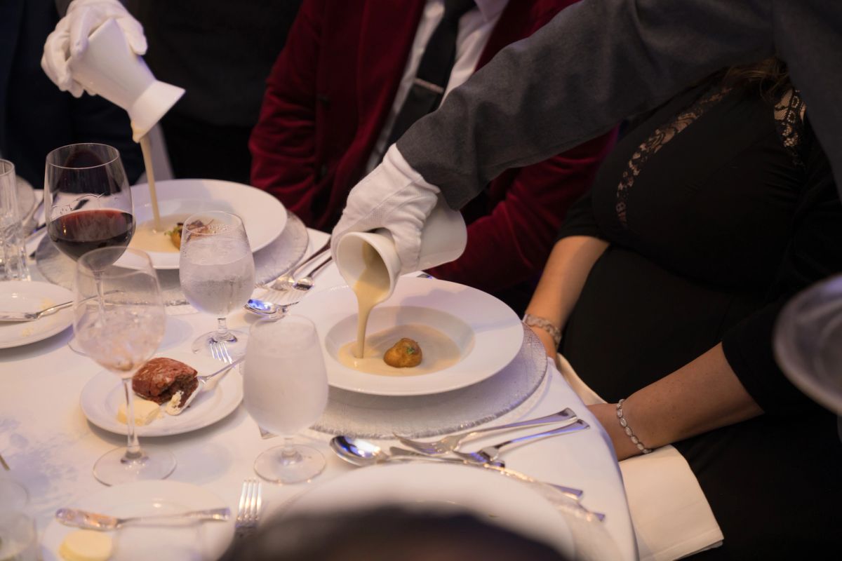 Server pouring sauce onto a plated dish at an elegant dinner table with wine glasses and formal place settings