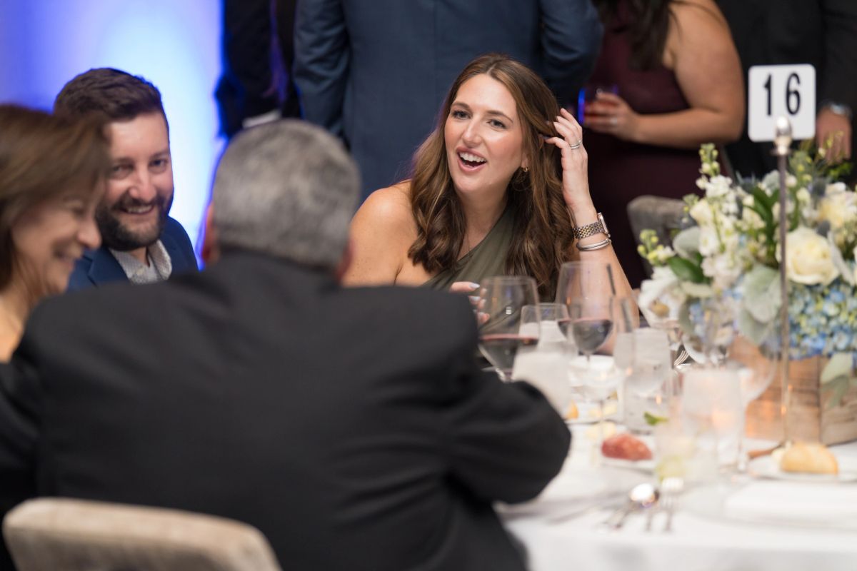 Woman laughing while seated at dinner table with other attendees at formal gala event with floral centerpiece