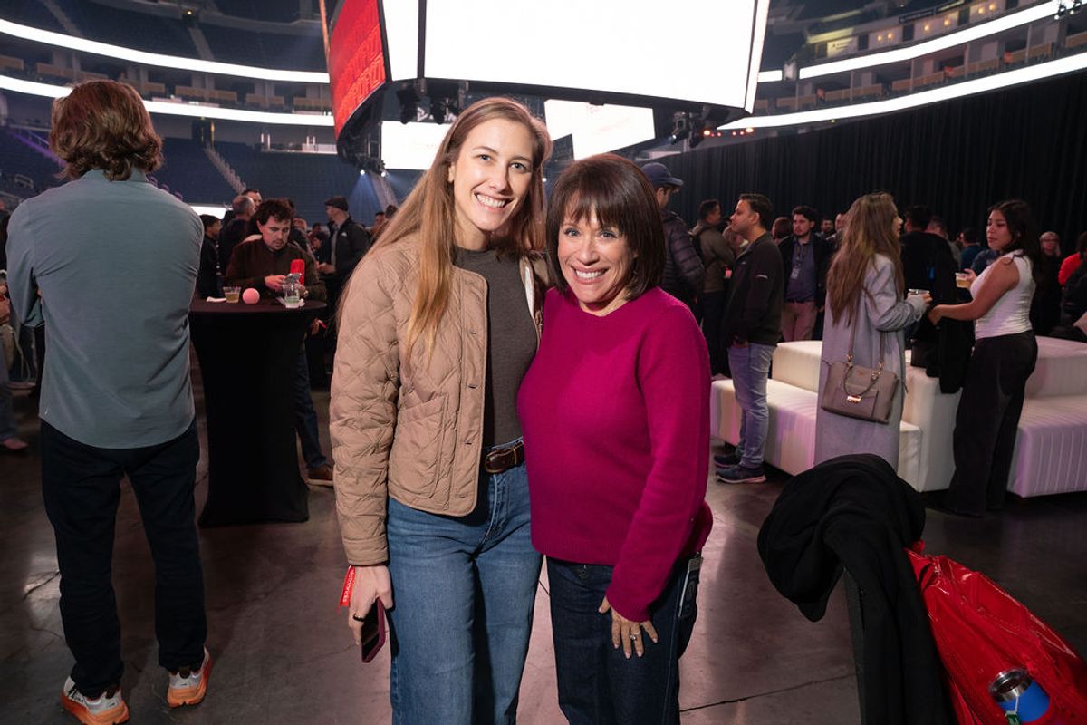 Two women smile together at a networking event in a modern venue with screens and attendees mingling