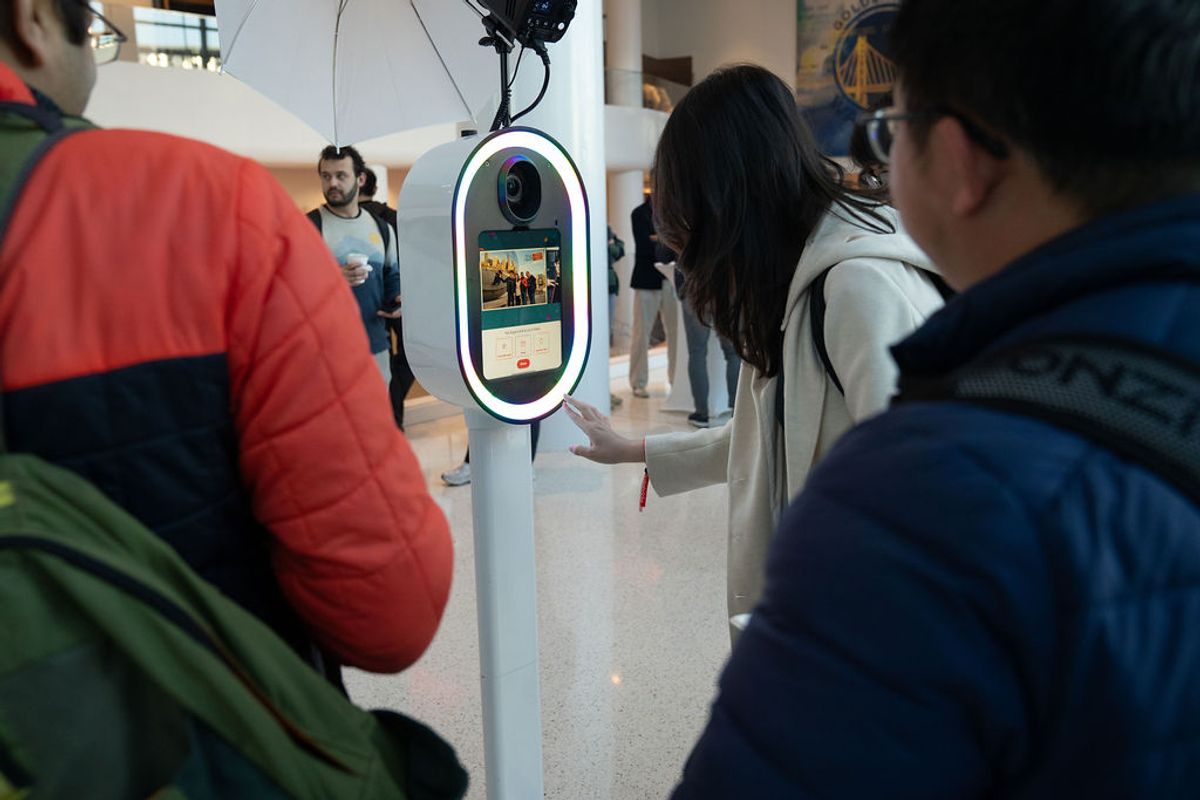 Conference attendees gather around an interactive kiosk with oval screen and camera at an indoor venue