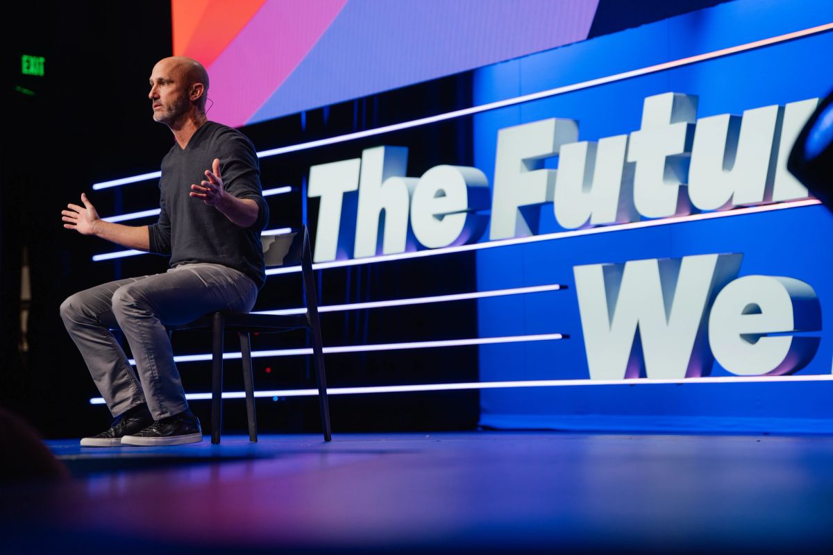 Speaker in black shirt presenting on stage with "The Future We" displayed on blue backdrop at conference venue