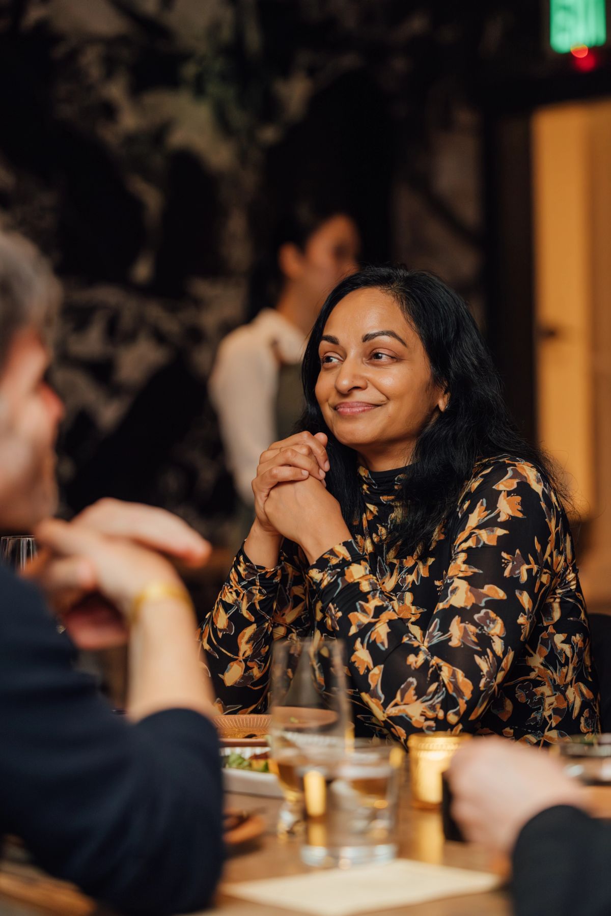 Woman in floral top smiling at dinner table with drinks and food, restaurant setting with blurred diners