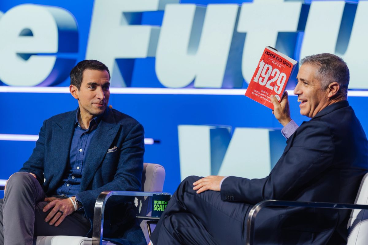 Two men in business attire sit in white chairs on stage, one holding up a red book during a conference.
