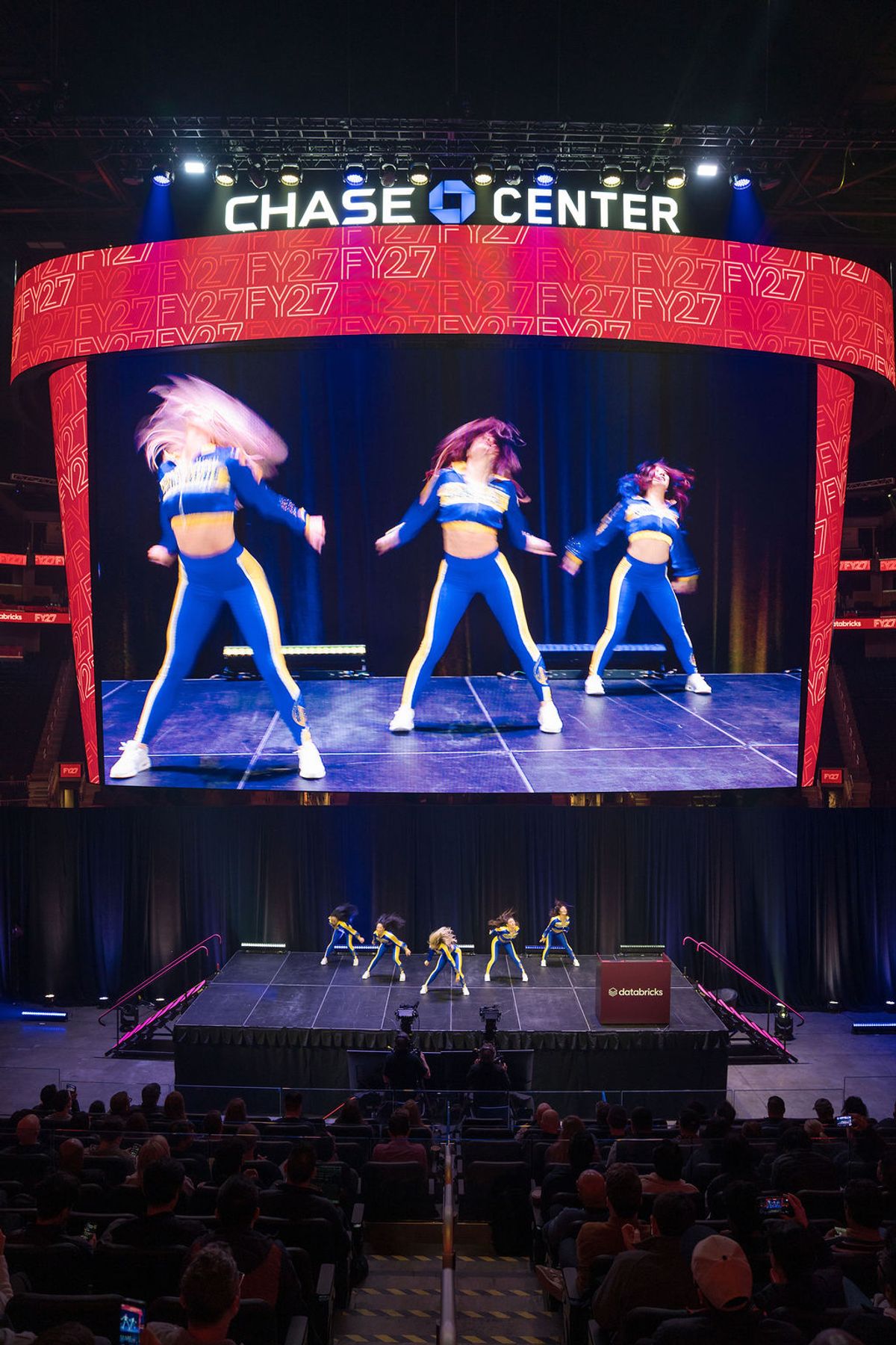 Performers on stage with cheerleaders displayed on large screen above at Chase Center during conference event