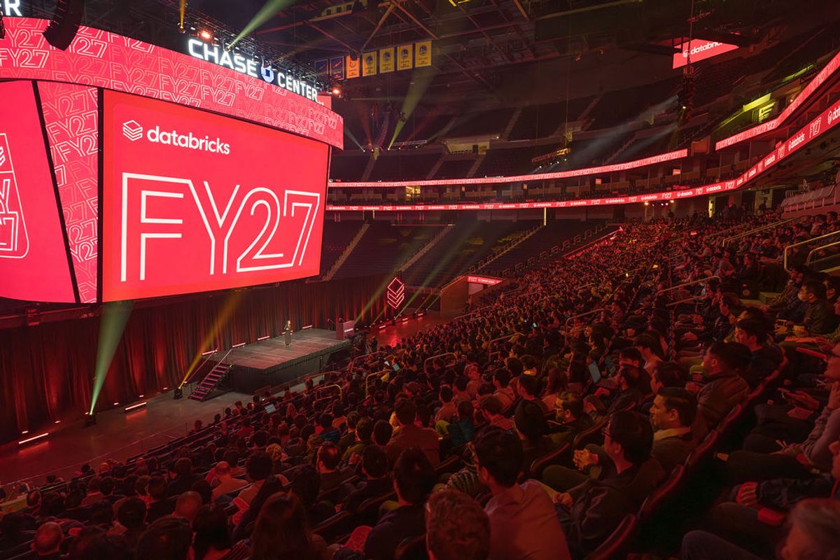 Large audience in arena seating facing stage with red lighting and "FY27" displayed on screens at Chase Center