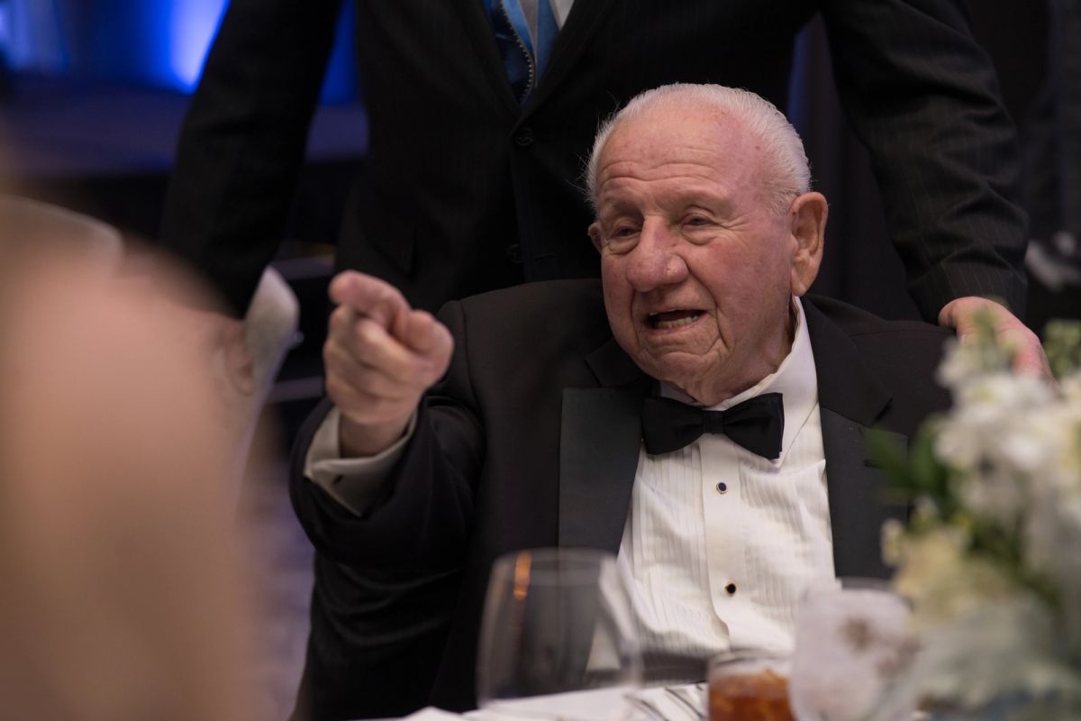 Elderly man in tuxedo gesturing while speaking at formal dinner table during gala event.