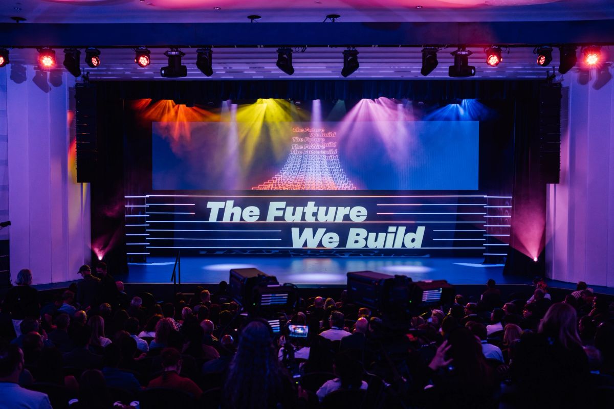 Conference stage with colorful spotlights and "The Future We Build" displayed on screen, audience seated below