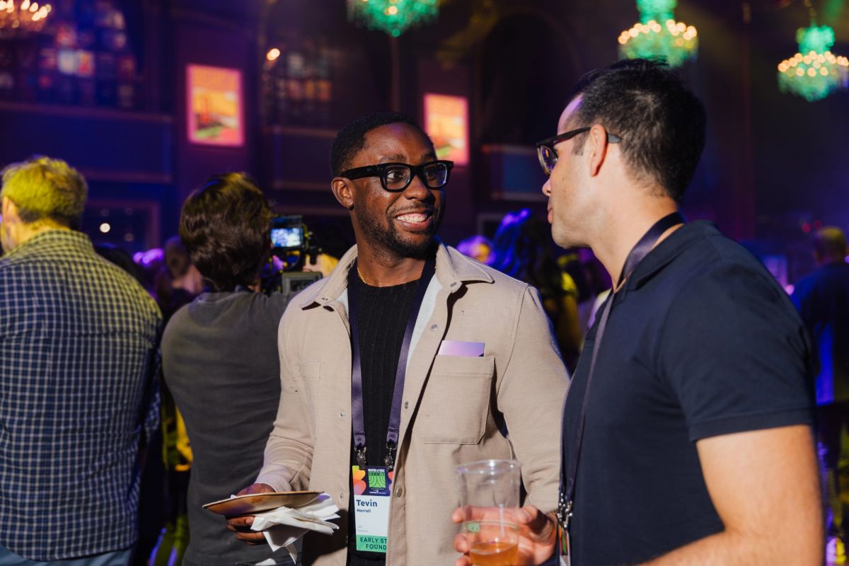 Two men in glasses having a conversation at a networking event, one holding a drink and papers