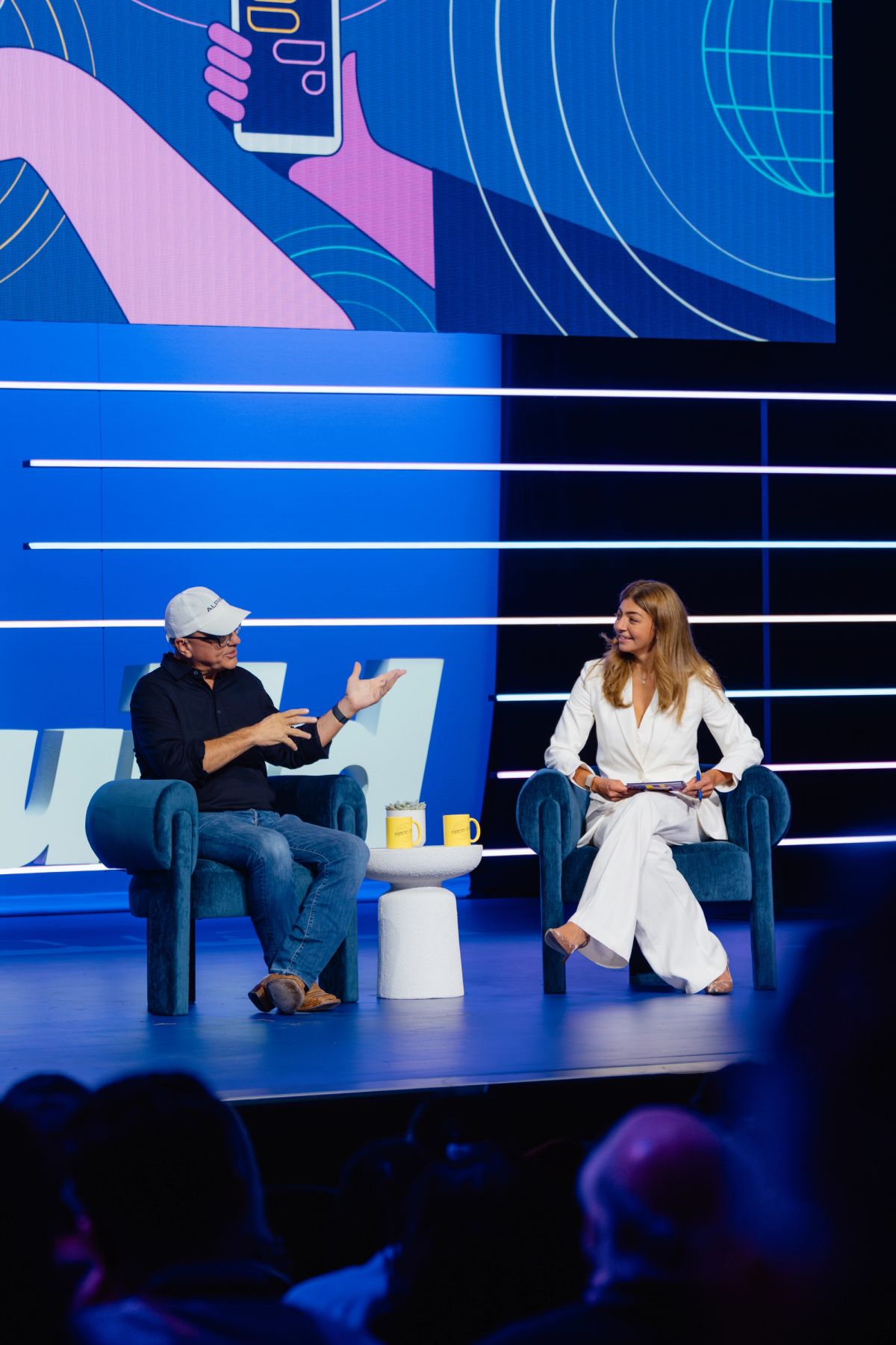 Two speakers in conversation on stage, man in casual wear and woman in white suit, seated in blue chairs with.