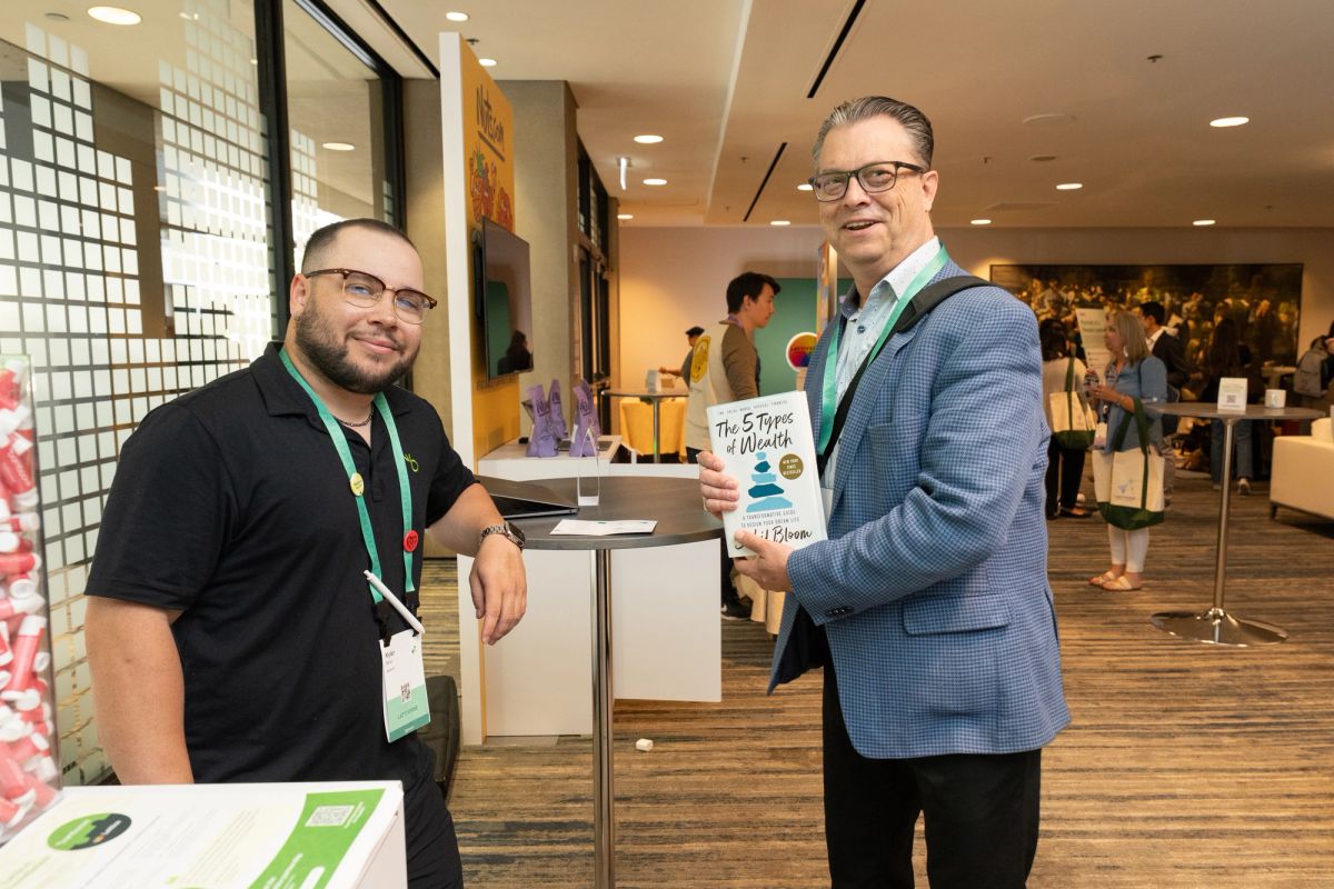 Two attendees smile holding "The 5 Types of Wealth" book at a conference networking area.