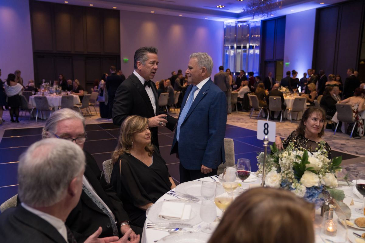 Two men in suits talking while standing at a formal dinner event with guests seated at decorated tables
