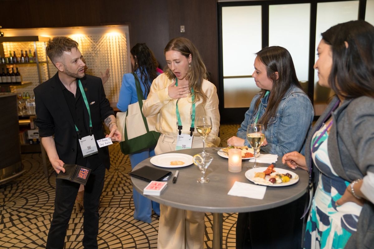 Attendees networking at a cocktail reception, reacting with surprise to a card magic trick.