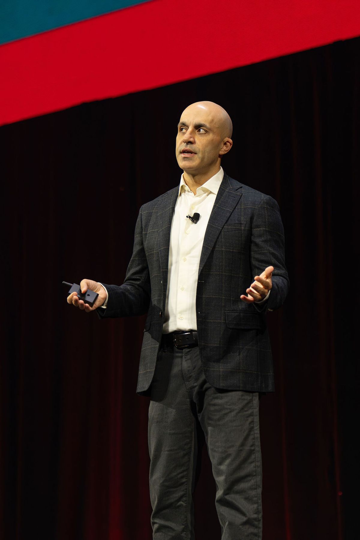 Speaker presenting on stage wearing gray suit and white shirt, holding device while gesturing