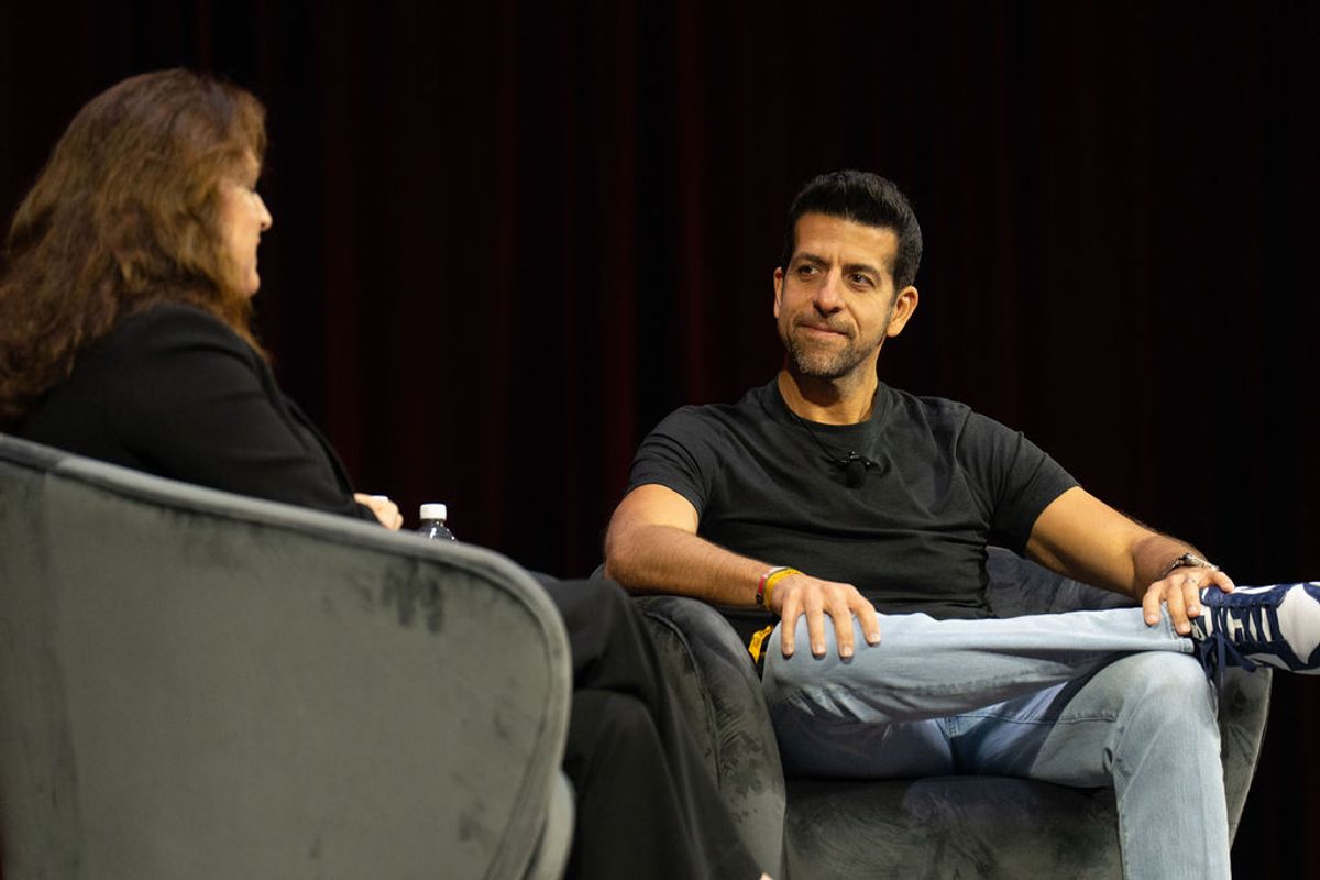 Two people in conversation on stage, one interviewer and one interviewee seated in chairs against dark.