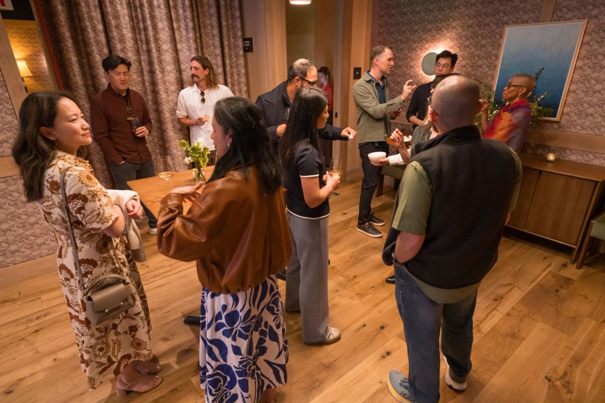 People networking and socializing in an indoor room with wood floors, patterned wallpaper, and curtains.