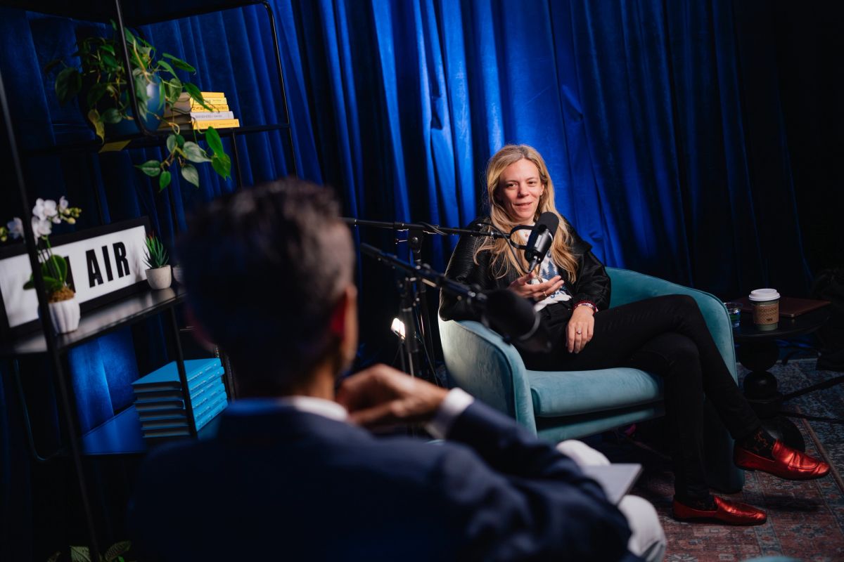 Woman speaking into microphone while seated in blue chair during interview, with interviewer visible in.