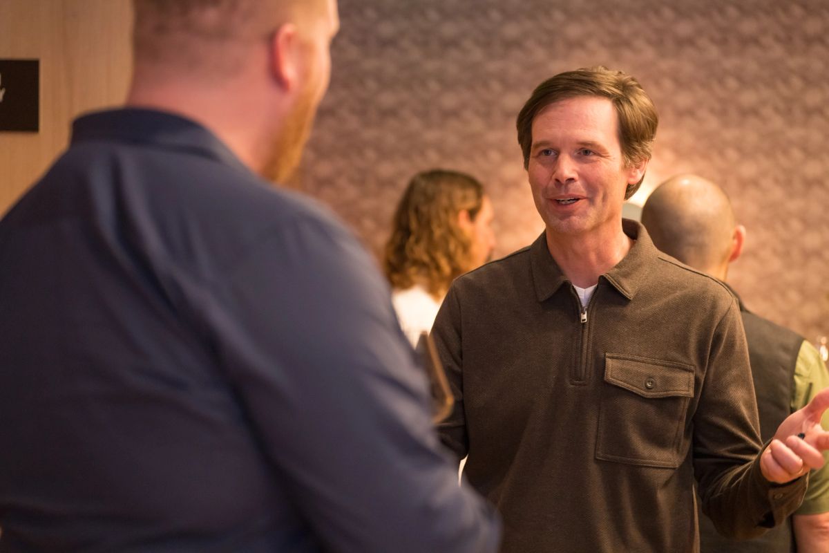 Two men in conversation at an indoor event, with other attendees visible in the background.