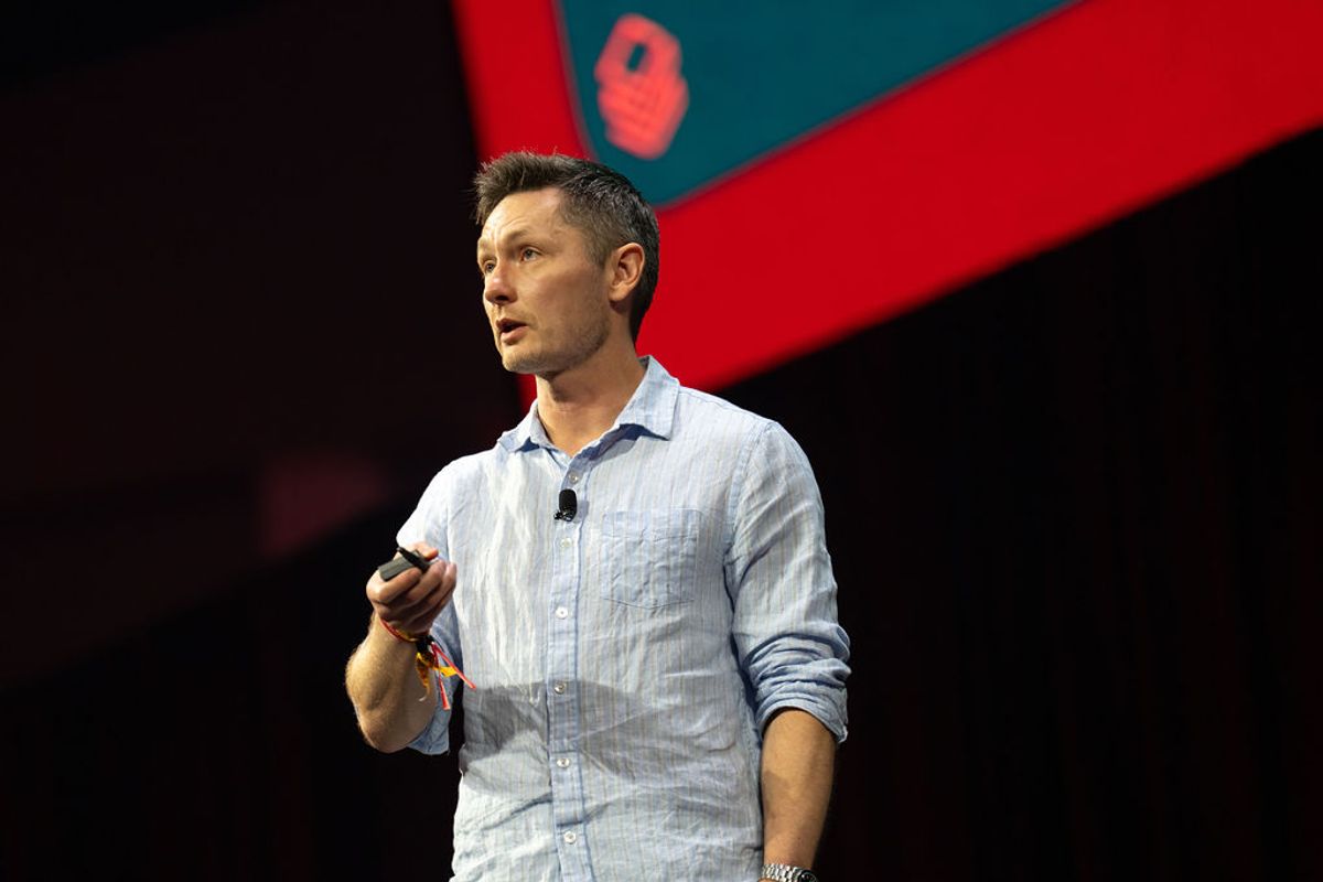 Speaker presenting on stage with microphone at conference event with red and teal backdrop design