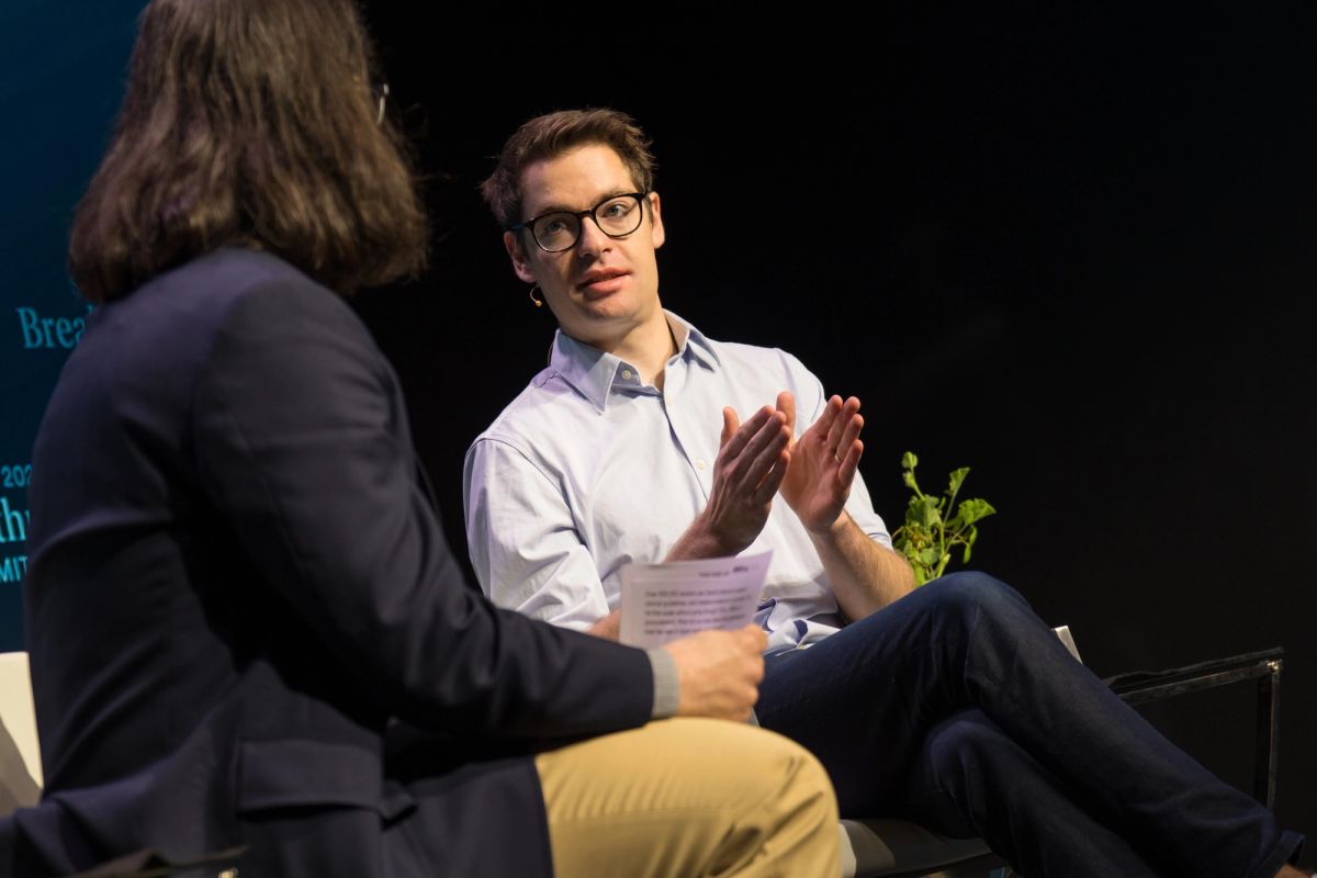 Two people in conversation on stage, one wearing glasses and light blue shirt gesturing while speaking to.