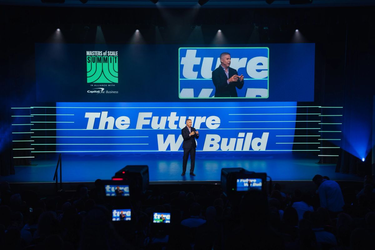 Speaker presenting on stage with "The Future We Build" backdrop and screens at conference venue